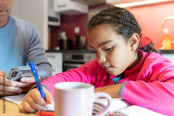 Fototapeta Young girl concentrating on school homework while learning, writing notes in a notebook at home, father nearby using a phone, combining education and modern family life