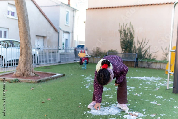 Fototapeta Young girl enjoying winter day playing with snow and ice on green artificial grass playground, experiencing childhood joy and curiosity
