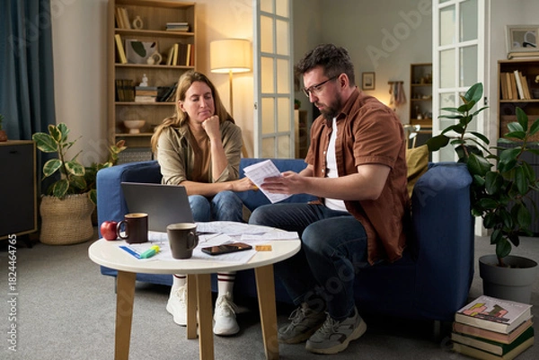 Obraz Middle aged Caucasian man explaining tax documents to middle aged Caucasian woman while sitting together at table with laptop and paperwork, both focused on financial planning