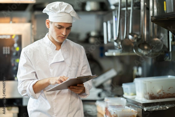 Obraz Young male chef in uniform with clipboard conducting inspection in restaurant kitchen