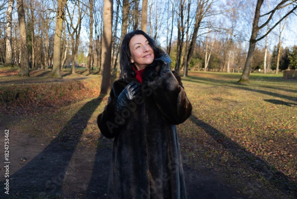 Fototapeta Mid-age woman in a luxurious fur coat smiles joyfully outdoors during winter, surrounded by trees and sunlight, capturing the essence of beauty and happiness in nature