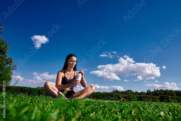Obraz Girl sits in Park on a background of clear sky and opens the water bottle.