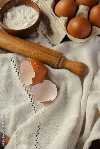 Fototapeta Vertical top view of freshly baked ingredients, a brown eggshell next to other whole eggs in a carton, bowls of flour and a rolling pin, ready to make homemade cakes and healthy recipes