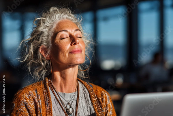 Obraz Woman enjoying a moment of relaxation in a sunlit cafe while working on a laptop, surrounded by a lively atmosphere