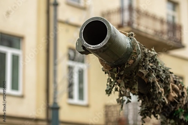 Fototapeta Close-up of a camouflaged tank cannon pointing towards the sky in an urban setting, with buildings, balconies and windows softly blurred in the background