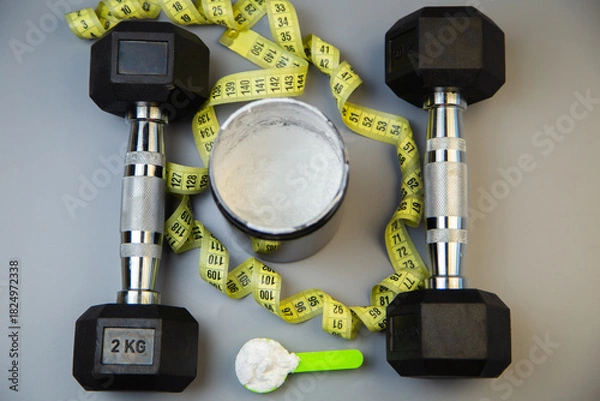 Fototapeta Black dumbbells are positioned alongside a measuring tape and a protein powder container, showcasing fitness essentials for a workout routine and healthy living