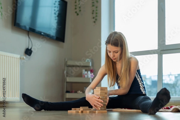 Fototapeta Teen girl stacking wooden blocks at home. A teenage girl sits on the floor with legs apart, carefully stacking wooden blocks during a quiet indoor activity in natural light.