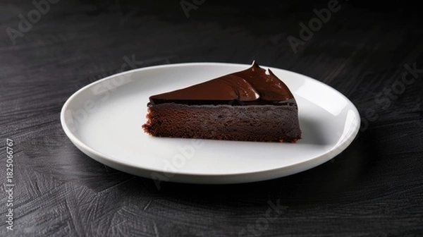 Fototapeta A triangular slice of rich, dark chocolate cake with a glossy, smooth glaze, presented on a minimalist white plate against a black, textured background