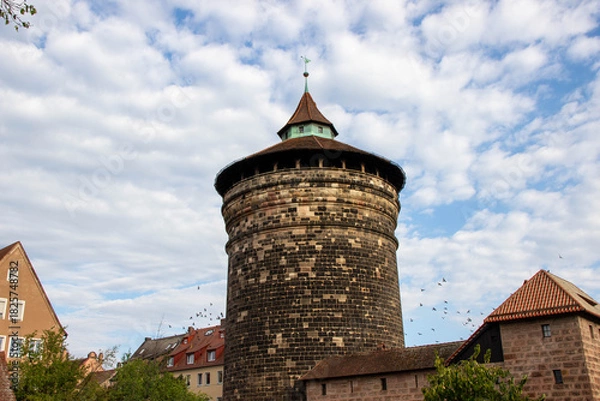 Fototapeta Historic stone tower with a conical roof stands tall against a blue sky filled with clouds, surrounded by traditional buildings and greenery, showcasing architectural heritage and urban landscape