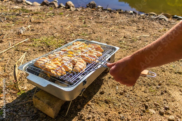 Fototapeta Chicken cooks on a portable grill by a lake during a hiking and outdoor travel stop. 