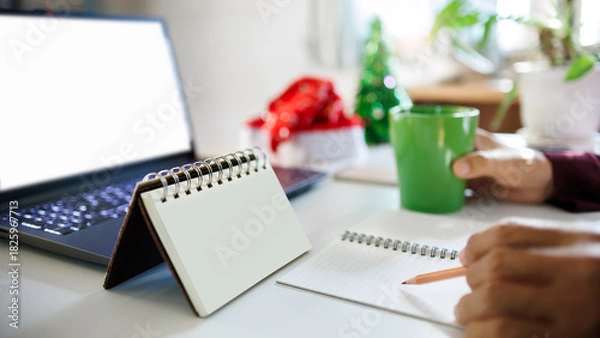 Obraz close up shot, hand holding pencil taking note on blank calendar working with laptop, red hat, christmas tree for decoration on desk, christmas holidays concept