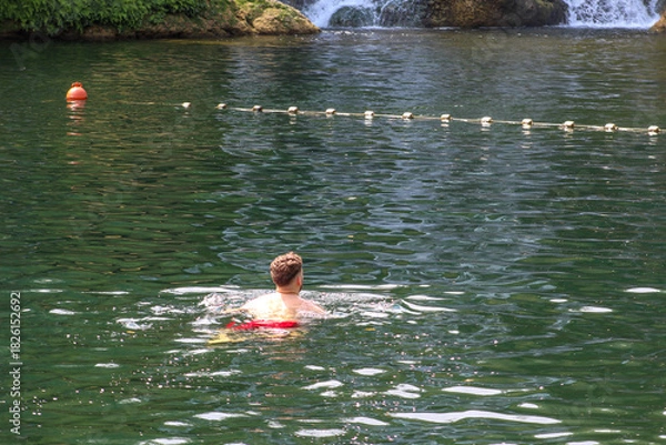 Obraz A man swimming in a clear green river near a small waterfall on a sunny day, surrounded by calm water and nature.