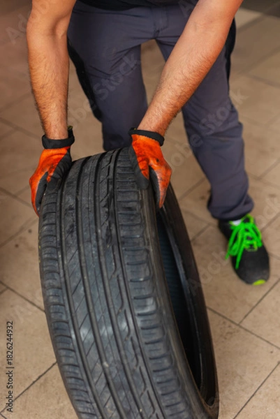 Fototapeta Mechanic wearing protective gloves rolling a wheel in a workshop for tire replacement. Professional car maintenance service and road safety.