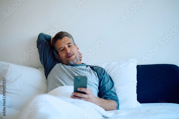 Fototapeta Man lying comfortably in bed, using his mobile phone, browsing applications, connected to technology in the bedroom