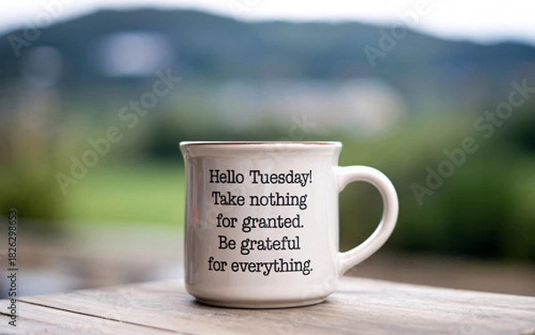 Fototapeta A white mug with a tuesday message sitting on a wooden surface with a blurred green background