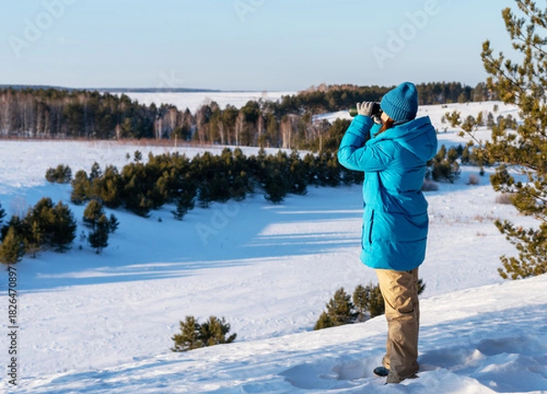 Fototapeta young woman in a blue jacket stands on a snowy landscape, using binoculars to observe the distant scenery of trees and fields, research in nature for ecological and conservation purposes, birdwatching