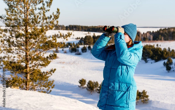 Fototapeta young Caucasian woman with wearing a blue winter coat and hat, uses binoculars in a snowy landscape with trees and hills in the background, Birdwatching and environmental research