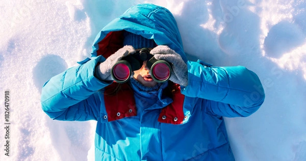 Fototapeta young woman wearing a blue winter jacket and gloves with surprised expression looks through binoculars while lying in snow. The scene captures a winter exploration theme