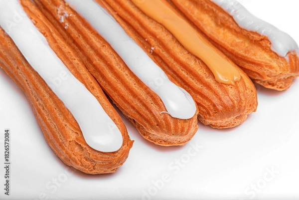 Fototapeta Four elongated choux pastries, eclairs, topped with white and caramel-colored icing on a bright white surface.