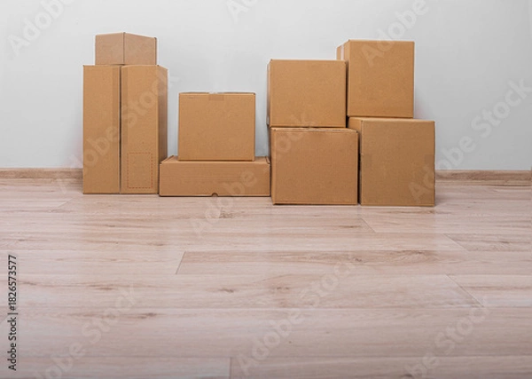 Fototapeta A collection of stacked, plain brown cardboard moving or shipping boxes rests on a light wood laminate floor against a white or light gray wall.