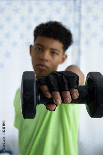 Obraz Brown skinned teenager in close up performing a front dumbbell raise, wearing gloves and training in the living room of his home. Concept of exercise and at home workout