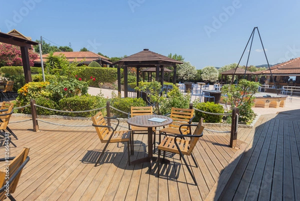 Obraz Wooden deck with table and chairs overlooking pool and gazebos in sunny resort garden.