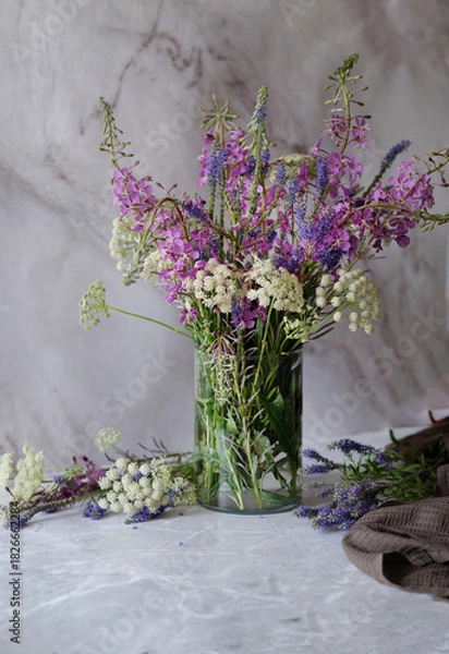 Fototapeta A vibrant bouquet of pink wildflowers is displayed in a clear glass jar on a gray surface, complemented by a piece of brown linen fabric draped nearby.