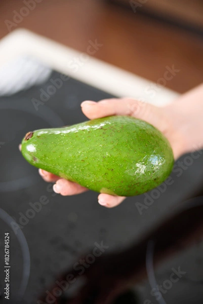 Fototapeta A large avocado in a woman's hand in the interiors of a bright and spacious kitchen