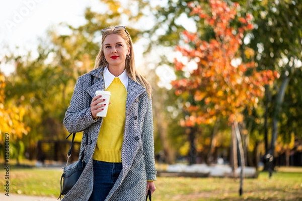Fototapeta Confident businesswoman in stylish autumn coat holding laptop bag and drinking coffee while walking through city park during sunny day.