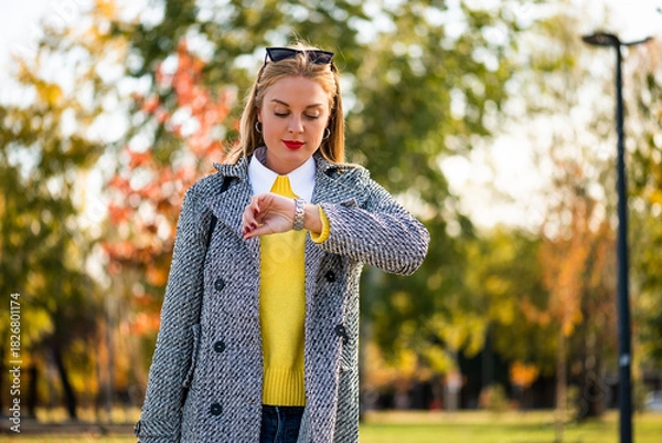 Fototapeta Stylish businesswoman with sunglasses in modern autumn coat using smartphone while walking through the city park during sunny day.