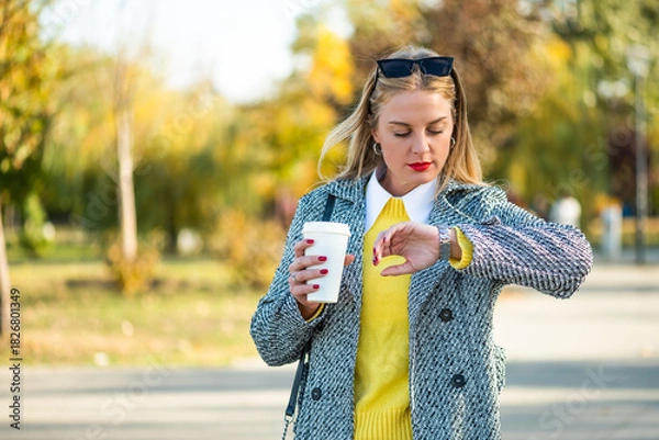 Fototapeta Confident businesswoman with sunglasses in stylish autumn coat checking the time on her wristwatch and drinking coffee while walking through the city park during sunny day.