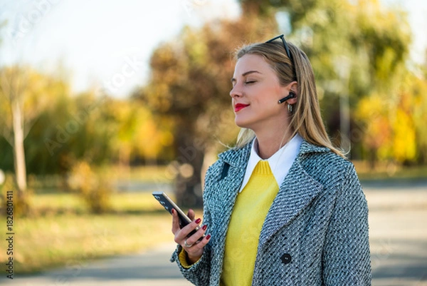 Obraz Close up portrait of beautiful urban businesswoman in autumn coat listening to music through earphones and using smartphone while standing in the city park on a sunny day.	