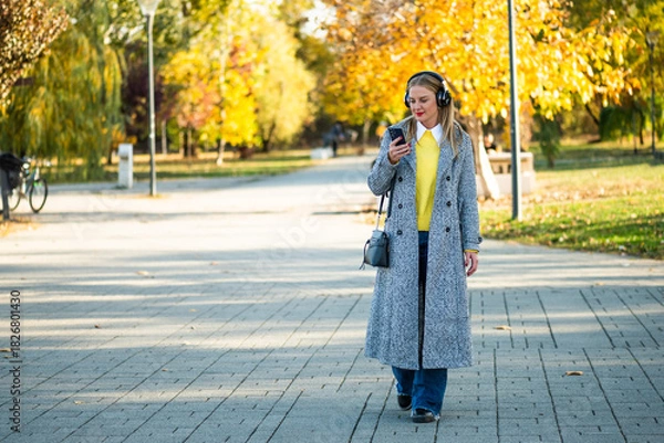 Fototapeta Urban businesswoman in stylish coat using wireless headphones and smartphone while walking through the city park during sunny autumn day.