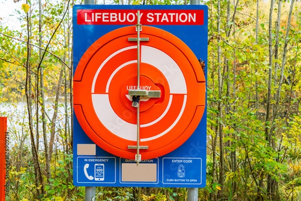 Fototapeta Lifebuoy Station containing a life saving ring or emergency life saver buoy at the edge of a lake, behind trees