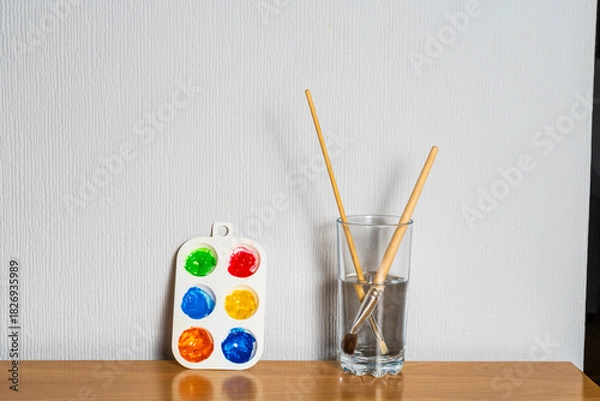 Fototapeta Paintbrushes in a jar of water with a palette of watercolour paints against a wall, minimal clean artist or hobby image