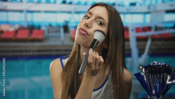 Fototapeta Woman in cheerleader uniform brushes cheek with makeup brush beside metallic pompom in building poolside area; confidence team ritual.