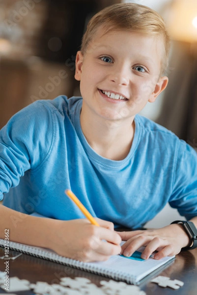 Obraz Mind development. Happy positive joyful boy writing in his copybook and smiling while doing his school task