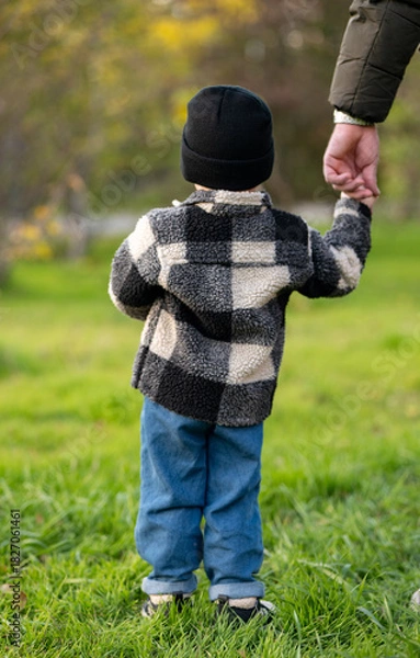 Fototapeta Young child holding an adult’s hand while standing on a grassy field. Dressed warmly, the child gazes ahead, evoking themes of care, safety, and connection in a natural outdoor setting.