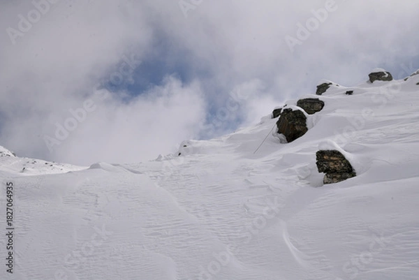 Obraz Snow-covered alpine slope with scattered rocks, strong wind patterns, and fog moving around a rugged mountain. Atmospheric winter landscape in cold high-altitude conditions.