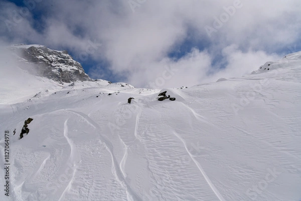 Obraz Snow-covered alpine slope with scattered rocks, strong wind patterns, and fog moving around a rugged mountain. Atmospheric winter landscape in cold high-altitude conditions.