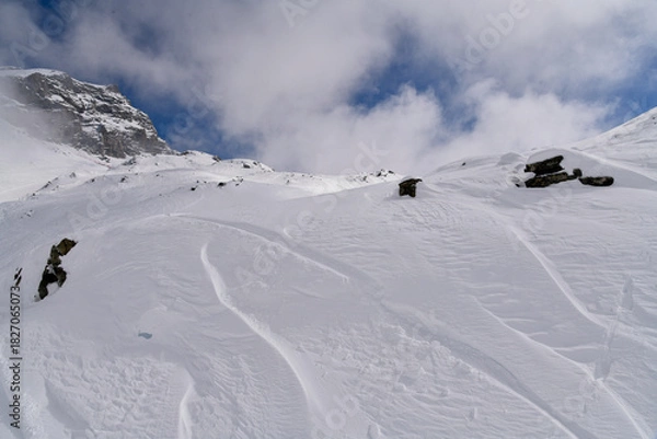 Obraz Snow-covered alpine slope with scattered rocks, strong wind patterns, and fog moving around a rugged mountain. Atmospheric winter landscape in cold high-altitude conditions.