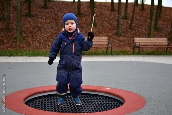 Fototapeta Toddler boy in a blue winter overall jumping on an in-ground playground trampoline. Behind him are park benches, autumn trees and soft rubber flooring on a cloudy day.