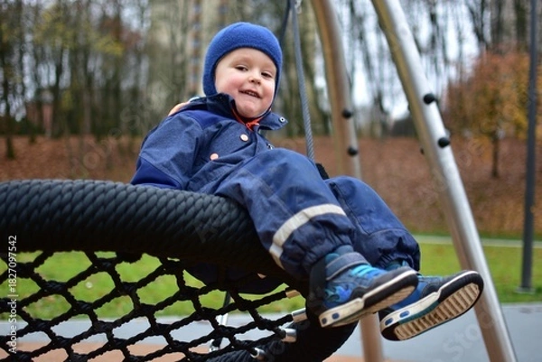 Fototapeta Smiling toddler boy in a blue winter suit sitting on a large rope swing at a modern playground. Autumn trees and soft play surfaces form a cozy outdoor scene.