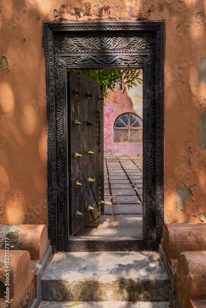 Obraz Ornate wooden door in an old orange wall opens to a sunlit courtyard.