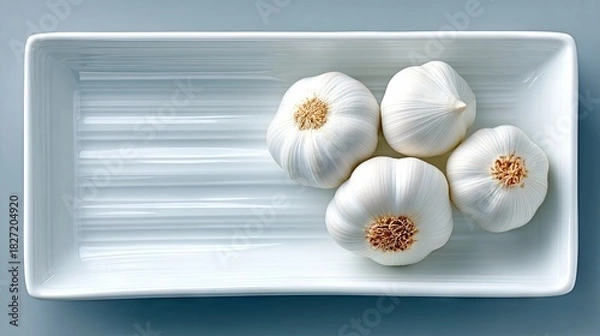 Fototapeta Four whole garlic bulbs are neatly arranged in a white, rectangular dish. The dish has a subtle ribbed texture on the inside. The background is a solid, muted b