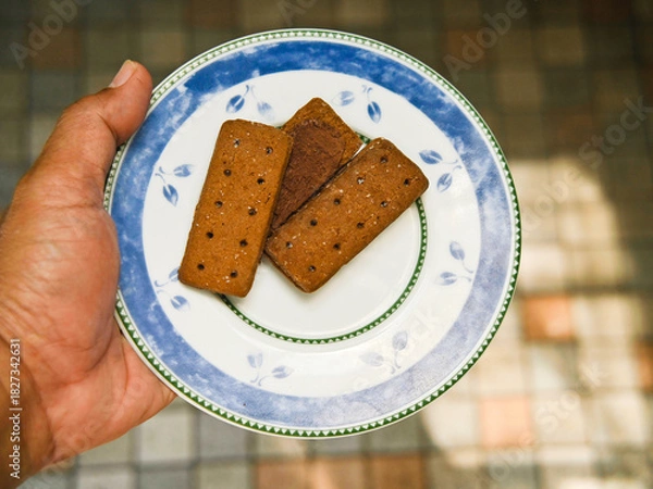 Fototapeta A man's hand carries a plate with several chocolate biscuits on it against a blurry ceramic tile background. Chocolate biscuits with granulated sugar for breakfast.