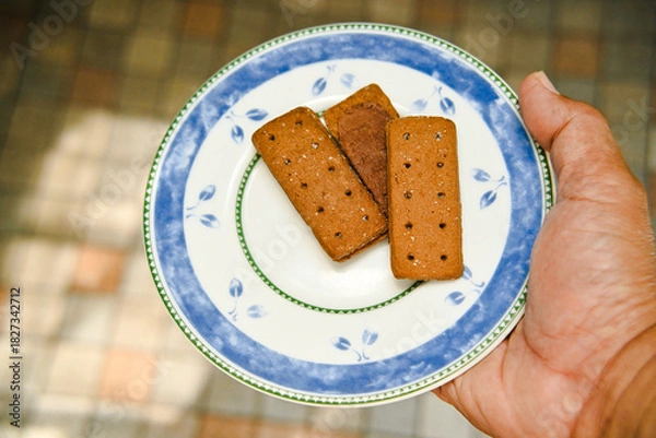 Fototapeta A man's hand carries a plate with several chocolate biscuits on it against a blurry ceramic tile background. Chocolate biscuits with granulated sugar for breakfast.