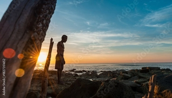 Fototapeta Silhouette of Man Standing on Rocky Beach at Sunset with Warm Colors and Dramatic Sky