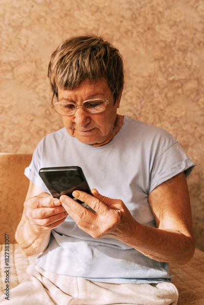 Obraz An elderly woman wearing glasses was intently immersed in her smartphone while sitting on a sofa in a sun-drenched. Digital Phone Addiction in Older Women, Screen Time Overload, and Modern Technology