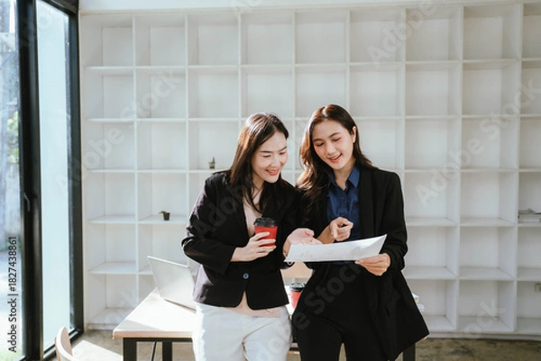 Fototapeta Two businesswomen work together at a laptop, smiling as they collaborate in an office with stacks of documents on the desk.